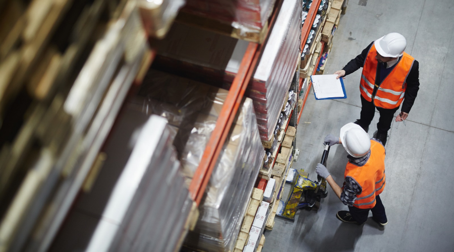 Two people in hard hats and reflective vests are at work in a warehouse setting, with shelves of stacked items stretching into the distance. One worker holds a clipboard and a pen, while the other operates a pallet jack to move materials.