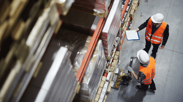 Two people in hard hats and reflective vests are at work in a warehouse setting, with shelves of stacked items stretching into the distance. One worker holds a clipboard and a pen, while the other operates a pallet jack to move materials.