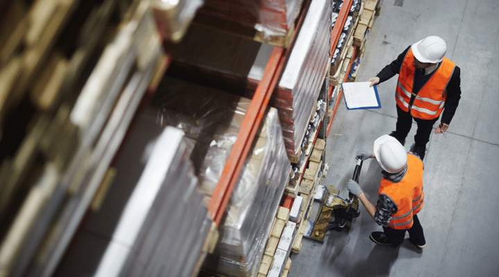 Two people in hard hats and reflective vests are at work in a warehouse setting, with shelves of stacked items stretching into the distance. One worker holds a clipboard and a pen, while the other operates a pallet jack to move materials.